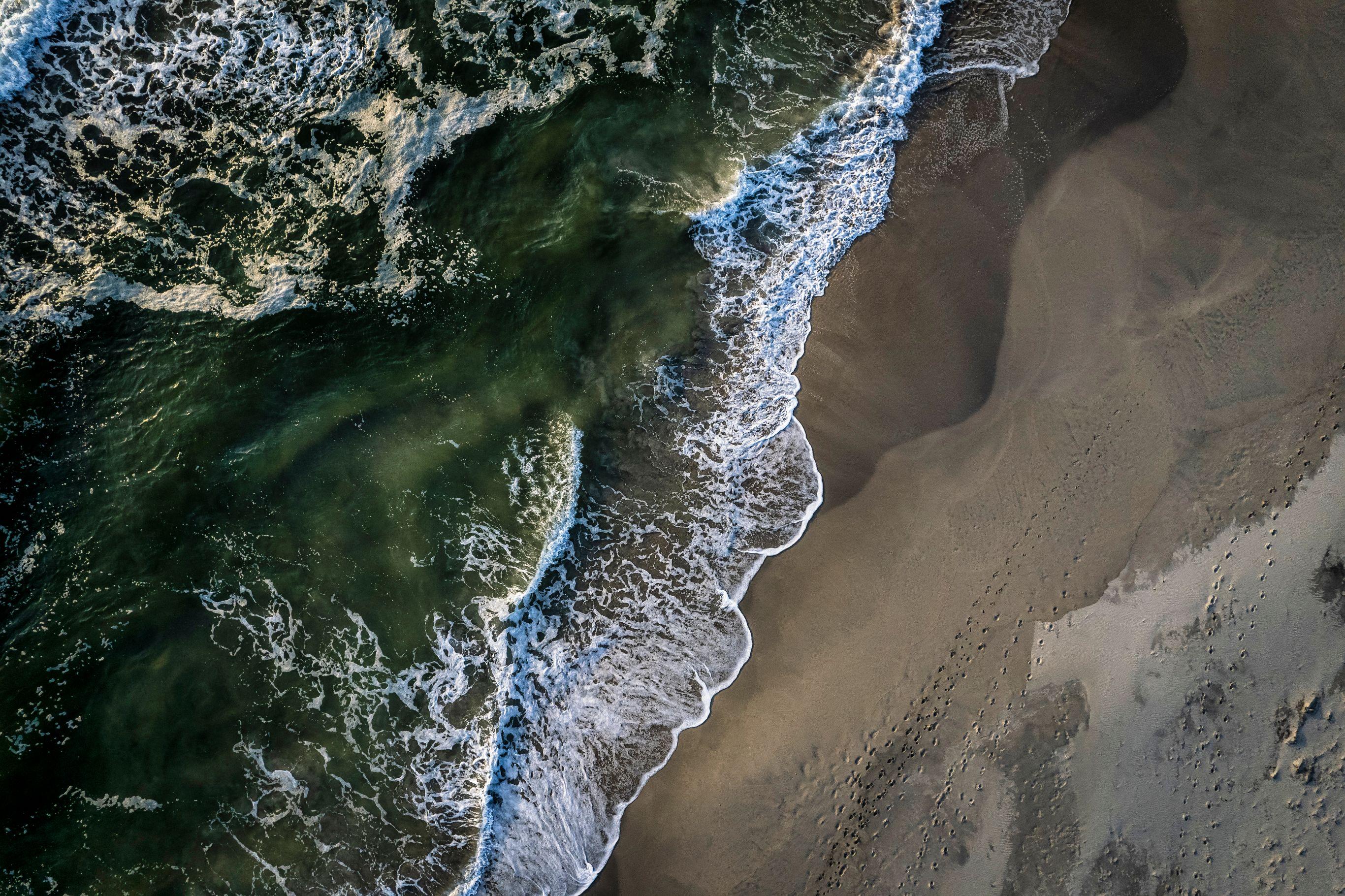 Directly overhead drone shot of a coastline where dark green ocean water meets a broad sandy beach. White foamy waves break diagonally across the frame, separating the deep water from the wet sand. Small footprints and bird tracks are visible in the dry sand on the right.
