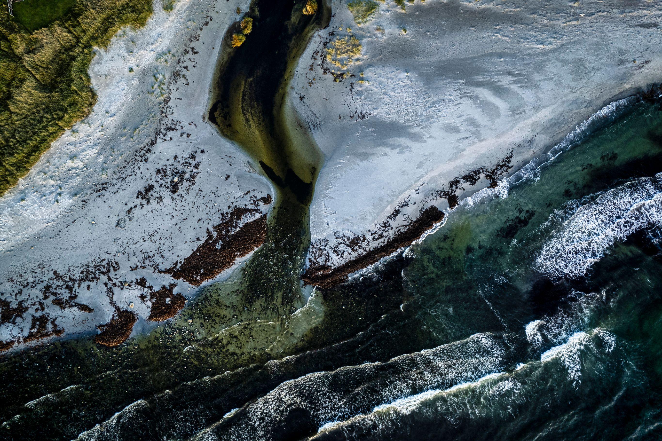 Overhead drone shot of a coastline where a narrow stream winds through a dusted beach before emptying into the sea. Dark brown seaweed and exposed rocks create jagged patterns across the shore. Turquoise ocean waves break in parallel lines on the right, while a strip of green vegetation and a few yellow-leafed trees are visible in the upper left corner.