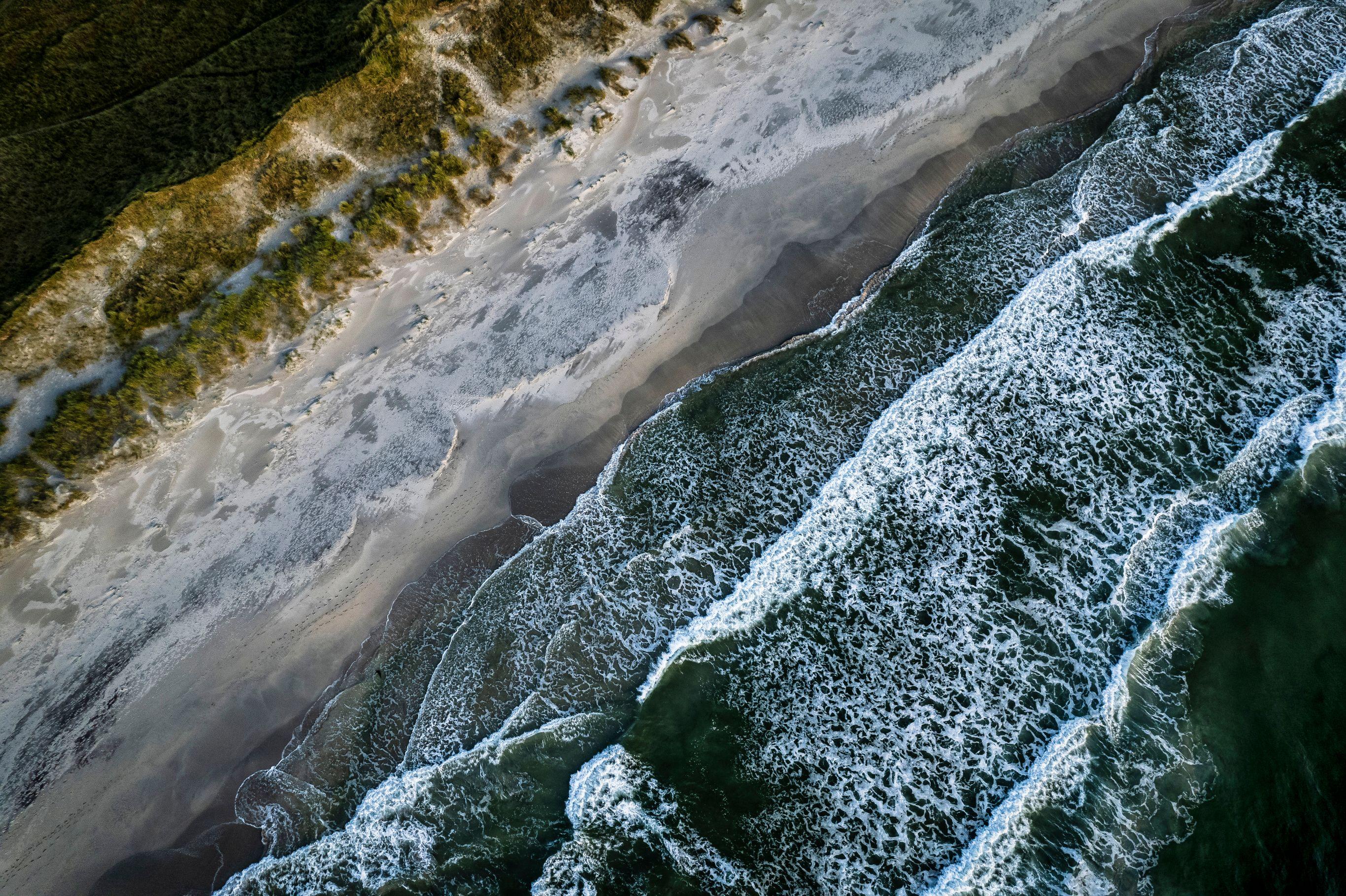 Overhead drone shot of a coastline divided diagonally between churning dark green ocean with white breaking waves on the right, a wide pale sandy beach in the middle, and a ridge of low vegetation-covered dunes in the upper left. Foam patterns left by receding waves mottle the wet sand.