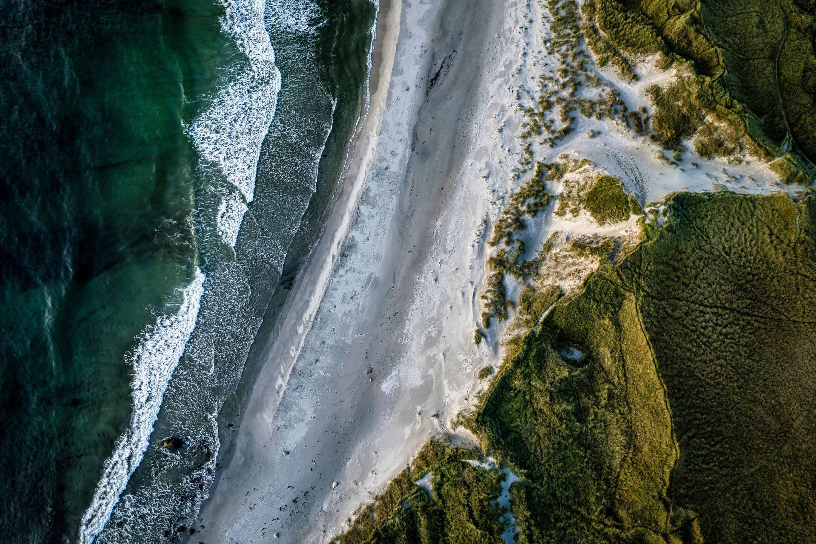 Overhead drone shot of a wide, deserted beach stretching vertically through the frame. Deep turquoise water with white breaking waves borders the left, while the right side is lined with textured green dune vegetation with patches of white snow or sand between the shrubs. A few scattered dark objects dot the shoreline.