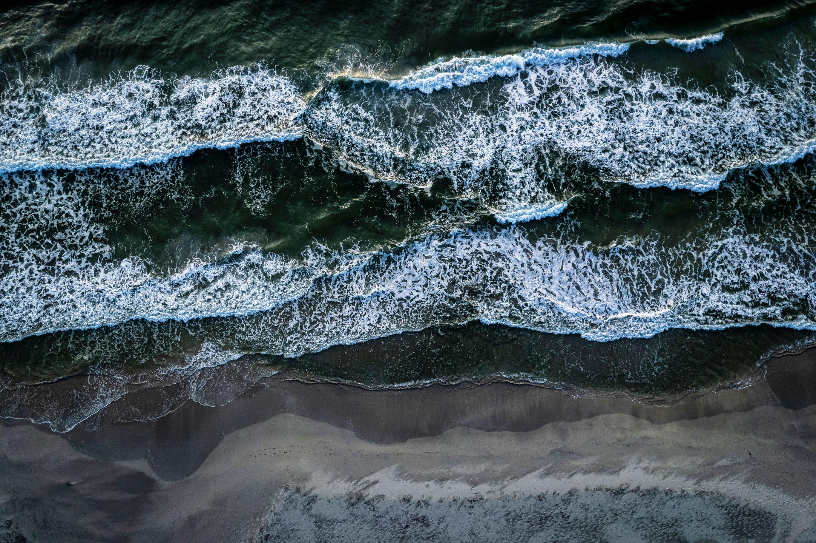 Directly overhead drone shot of a shoreline with three distinct wave lines rolling in parallel toward a wide, dark sandy beach. The white foam of each breaking wave contrasts sharply against the deep dark green ocean. The lower third of the frame shows wet sand with subtle ripple patterns left by the receding water.